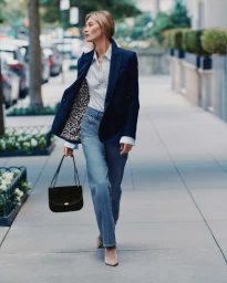 Grace Elizabeth walking on a city street wearing a navy blazer, striped shirt, and blue jeans holding a black handbag.