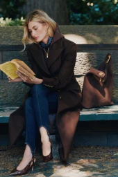 Grace Elizabeth reading a book on a stone bench wearing a dark brown coat and blue stirrup pants for Veronica Beard Fall 2026