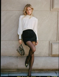 Grace Elizabeth leaning against a stone wall in a white blouse, black skirt, sheer tights, holding a zebra-print handbag