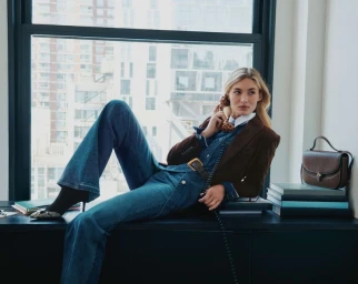 Grace Elizabeth seated on a windowsill holding a rotary phone wearing denim flare jeans and a brown blazer for Veronica Beard fall campaign