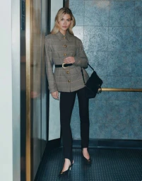 Grace Elizabeth leaning against an elevator door wearing a plaid jacket and black trousers for Veronica Beard campaign.