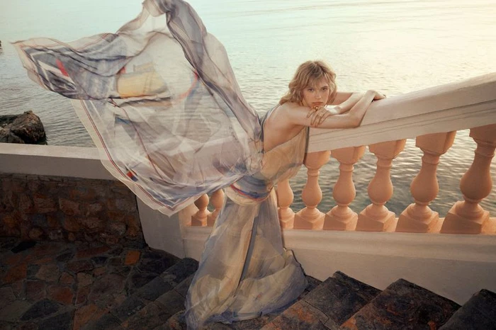 Model leaning on a balustrade near the sea wearing a flowing patterned dress with a large billowing fabric piece