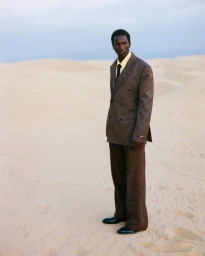 Black man standing in sandy dunes wearing a brown double-breasted suit and yellow shirt for At Large Magazine.