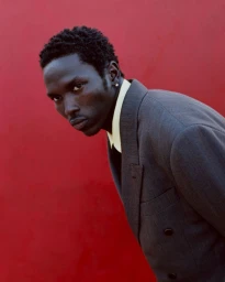 Black male model wearing a grey blazer and yellow shirt against a red wall for At Large Magazine