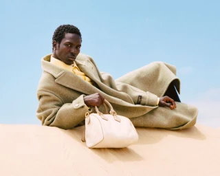 Model Monykuoch Deng lying on sand dunes wearing a heavy coat and holding a light handbag against a clear blue sky