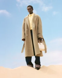 Model Monykuoch Deng standing on a sandy dune wearing a long camel coat and yellow shirt against a clear blue sky