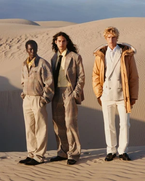Three models standing on sand dunes wearing tailored suits, jackets, and earth-toned trousers under a clear sky.