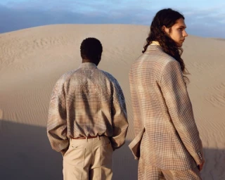 Two models standing in a sand dune landscape, one wearing a patterned blazer and the other a shimmering sequined top.