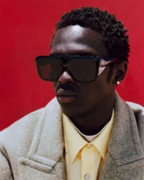 Portrait of Monykuoch Deng wearing oversized dark sunglasses, a yellow shirt, and a grey textured coat against a solid red background