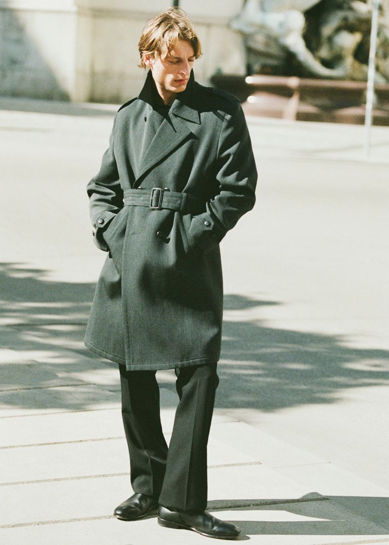 Male model wearing a dark belted coat and trousers standing outdoors in sunlight for Le Figaro editorial.