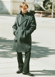 Male model wearing a dark belted coat and trousers standing outdoors in sunlight for Le Figaro editorial.
