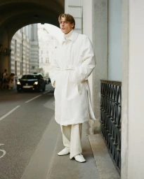 Model Colin Otto wearing a long white belted coat and matching trousers standing on a city street for Le Figaro.