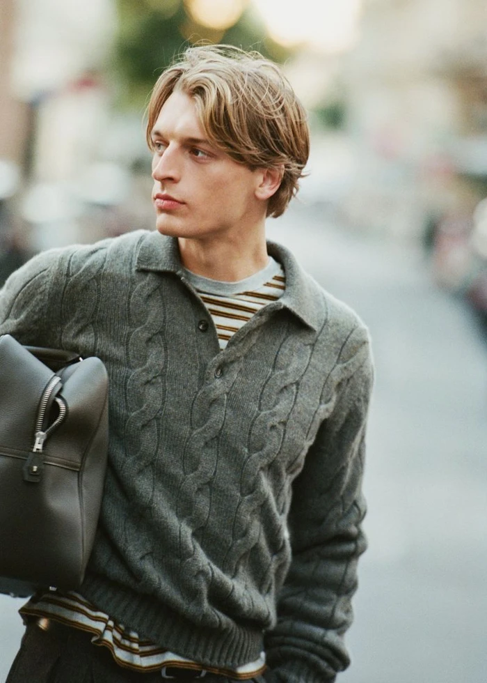 Model Colin Otto wearing a cable knit polo sweater and carrying a dark bag outdoors for Le Figaro, photographed by Valentin Hennequin.