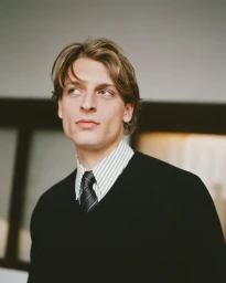 Portrait of a man wearing a black sweater over a striped button-down shirt and tie, looking off to the side.