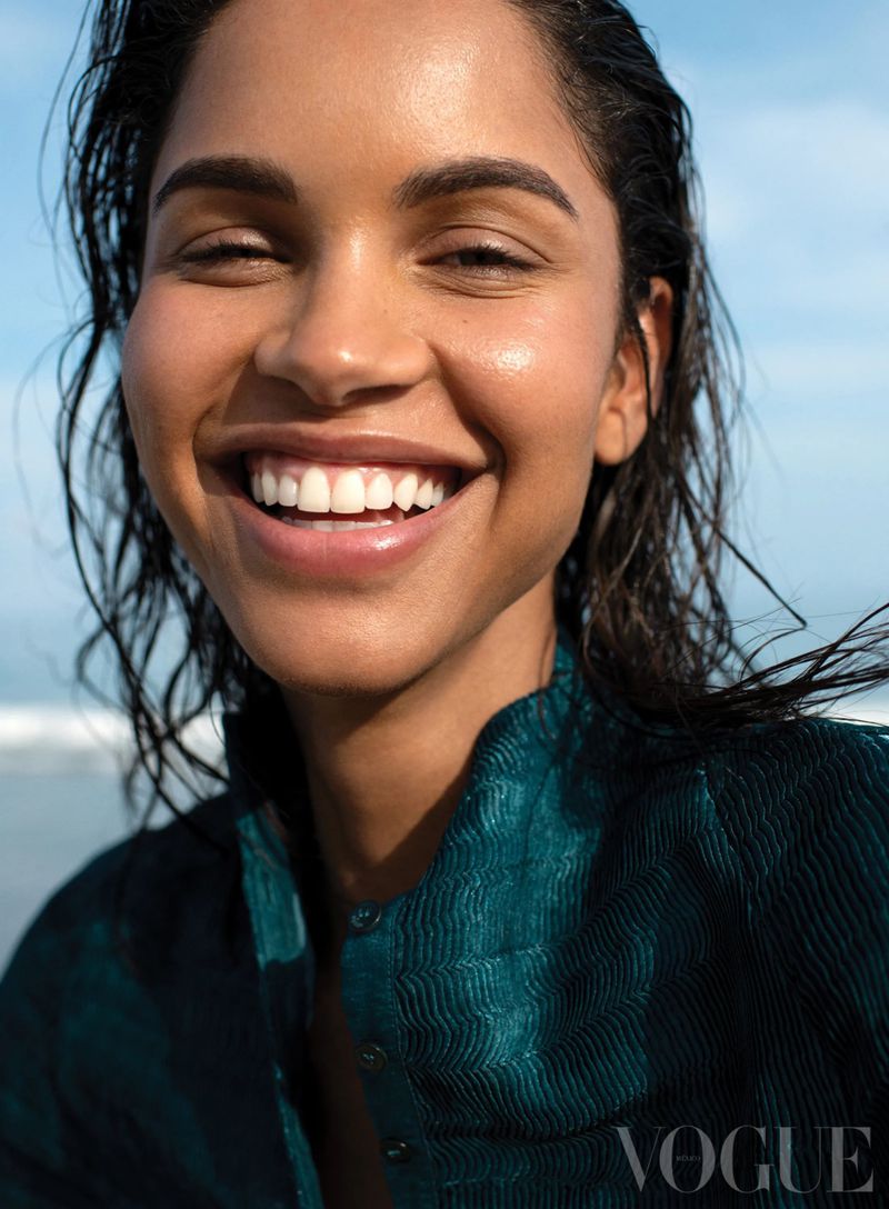 Close-up of a smiling adult female with wet dark hair, wearing a teal top, against a backdrop of the sky and ocean.