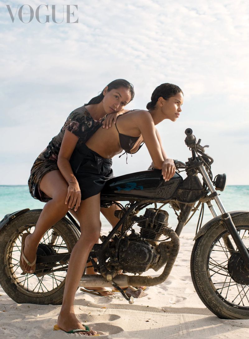 Two adult females pose on a motorbike on a sandy beach near the ocean under a bright sky; Vogue logo visible in the corner.