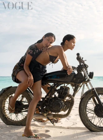 Two adult females pose on a motorbike on a sandy beach near the ocean under a bright sky; Vogue logo visible in the corner.