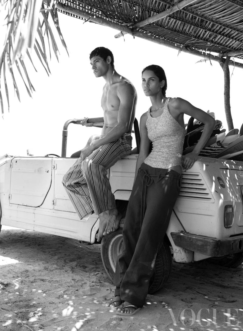 A shirtless man and woman in casual pants leaning on a white car with surfboards on the roof, shot in black and white with a beach background.