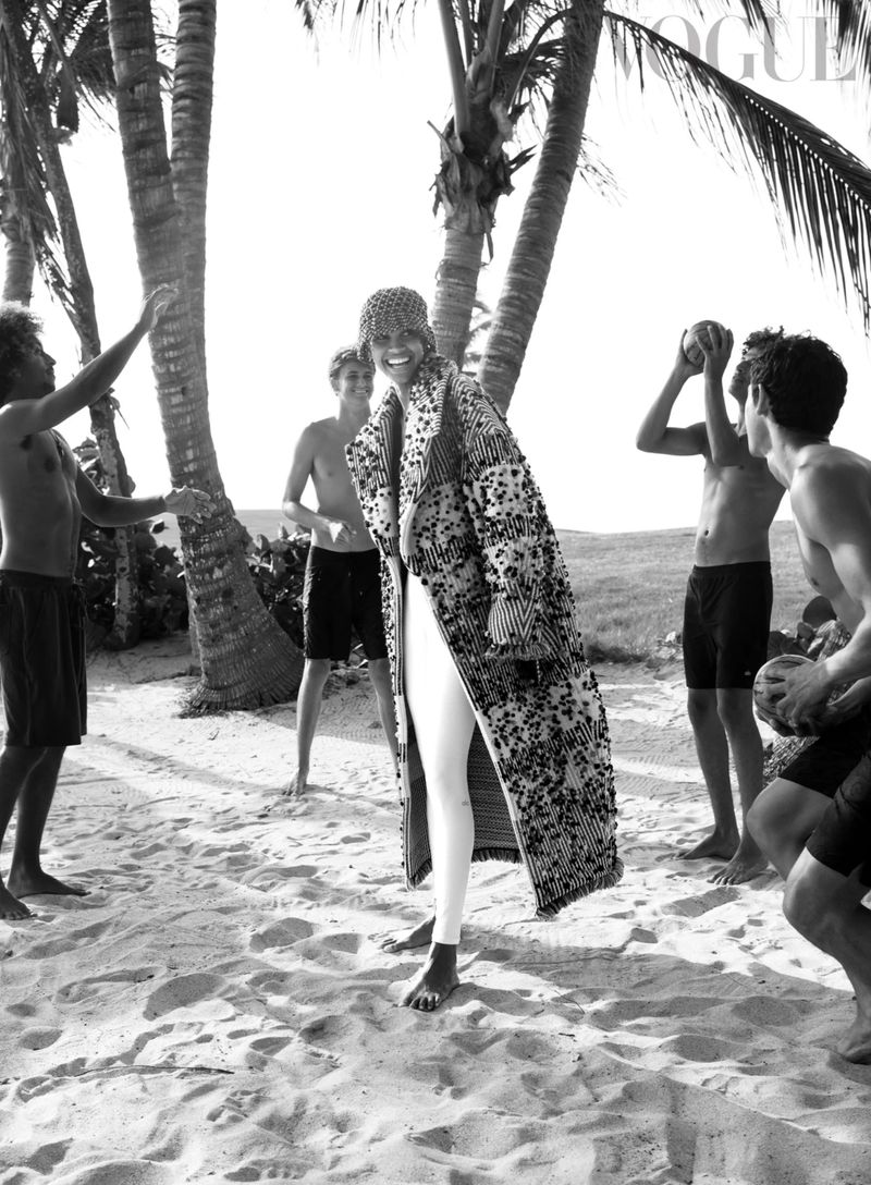 Black and white fashion editorial featuring a woman in a patterned coat posing on a beach with several young men and palm trees.