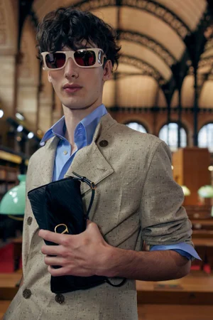 A young male model with dark curly hair wearing off-white sunglasses, a blue collared shirt, and a beige blazer while holding a black clutch.
