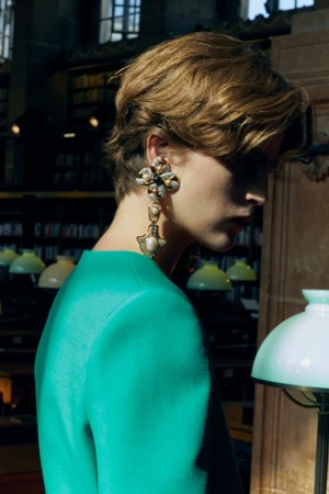 Model wearing a vibrant turquoise top and large jewelled dangling earring in an interior library setting.