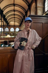 Model wearing a pink trench coat, blue baseball cap, and holding a black handbag inside a historic library interior
