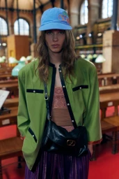 Model wearing a blue cap, green jacket, and black crossbody bag inside a library setting