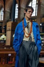 Model in vibrant blue coat and striped tie wearing amber bead necklaces inside a grand historic library interior