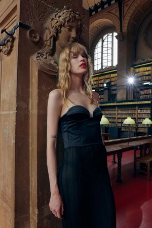 Model in black strapless corset top leaning against carved stone pillar in a library with wooden bookshelves