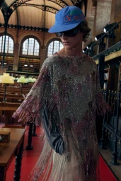 Model wearing a blue baseball cap and embellished fringe tunic inside a large historic library with wooden desks.