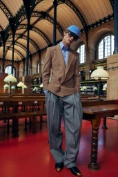 Model wearing a brown double-breasted blazer, light blue shirt, grey pleated trousers, sunglasses, and a blue cap inside a historic library.