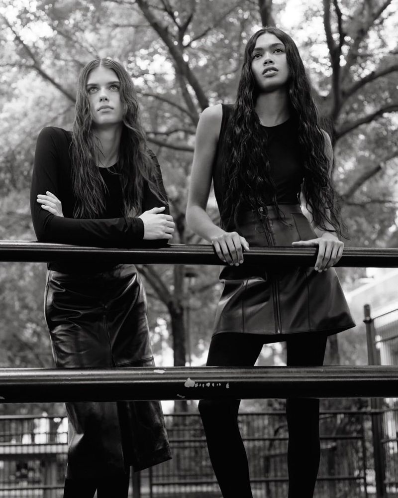 Two female models in black outfits leaning against a metal railing outdoors in a monochromatic fashion editorial photograph.