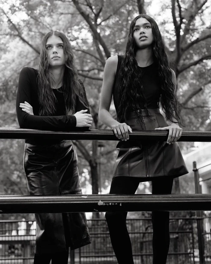 Two female models in black outfits leaning against a metal railing outdoors in a monochromatic fashion editorial photograph.