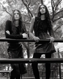 Two female models in black outfits leaning against a metal railing outdoors in a monochromatic fashion editorial photograph.