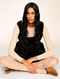 Model Thalita Ferreira with long dark hair wearing a sleeveless brown fur dress over a beige top against a white seamless background