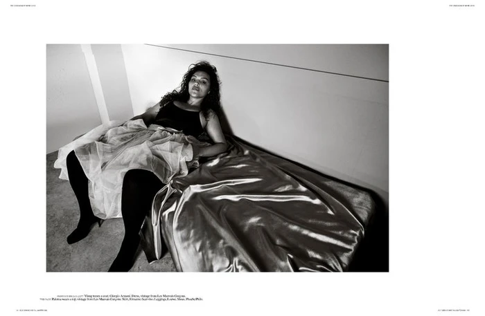Model reclining on a shiny satin bed wearing a dark top and layered skirt with tights