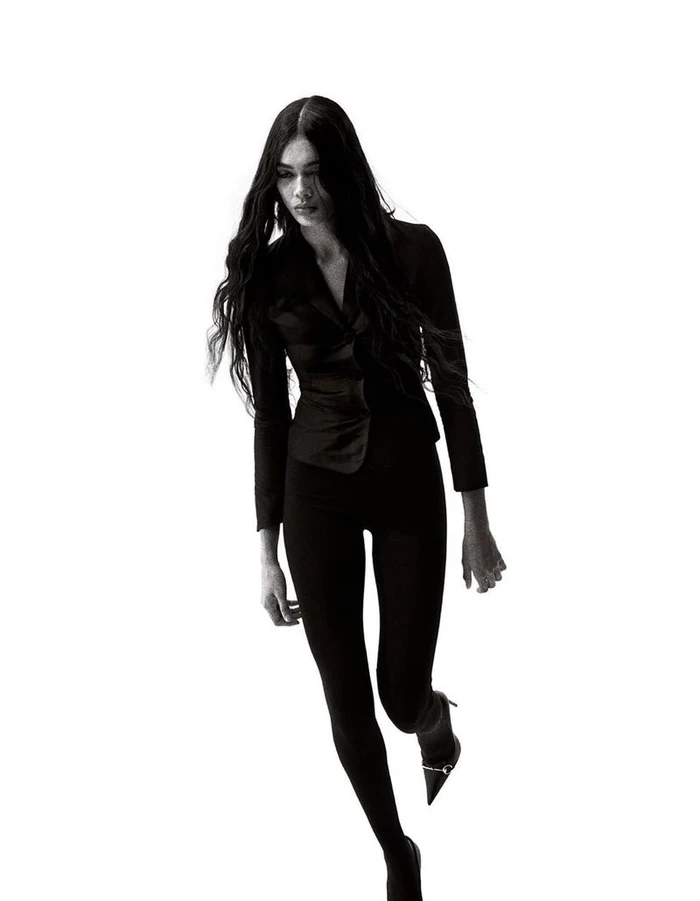 Model with long dark wavy hair wearing a black jacket and leggings against a white background