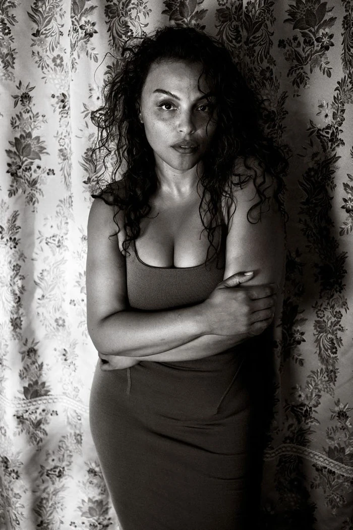Portrait of a woman with dark curly hair wearing a dark fitted dress against a patterned floral curtain background.