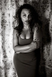 Portrait of a woman with dark curly hair wearing a dark fitted dress against a patterned floral curtain background.