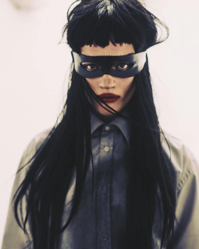 Model with long black hair and heavy bangs wearing a leather eye mask and grey buttoned shirt against a white background