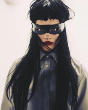 Model with long black hair and heavy bangs wearing a leather eye mask and grey buttoned shirt against a white background