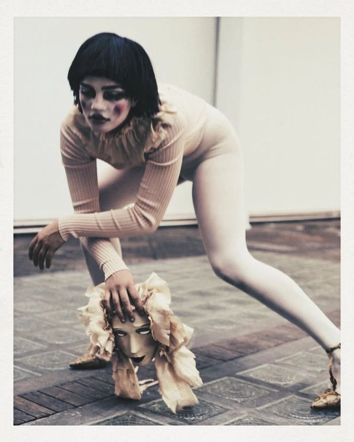Model in cream bodysuit and dark bob wig holding a decorative theatrical mask on a tiled floor