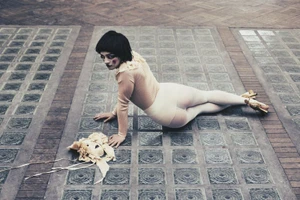 A woman wearing a beige top and white tights lying on a patterned glass brick floor, looking back at the camera.