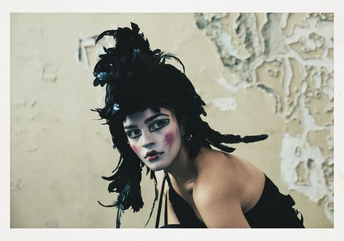 Model with dramatic theatrical makeup and a black feathered headdress against a weathered wall background
