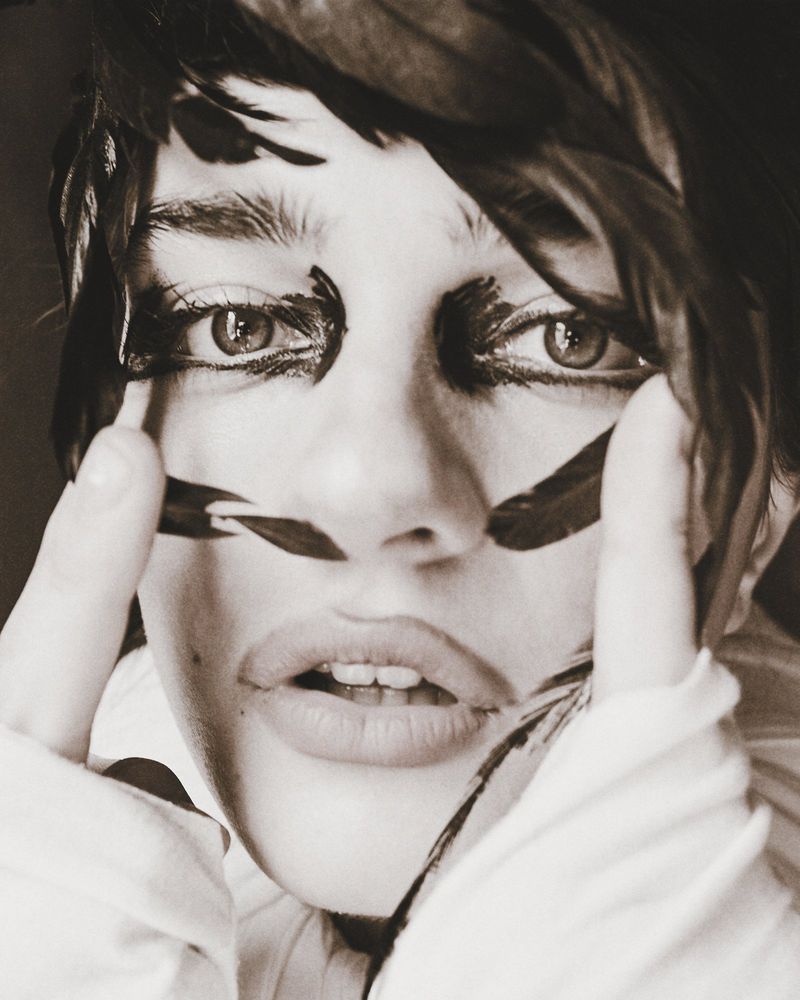 Close-up portrait of a model with dark feathers framing her face and bold black eye makeup for SHADOWPLAY Magazine