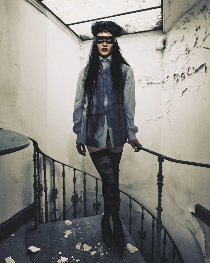 Model wearing a black eye mask, oversized button-down shirt, and patterned thigh-high stockings on an interior spiral staircase.