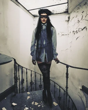 Model wearing a black eye mask, oversized button-down shirt, and patterned thigh-high stockings on an interior spiral staircase.