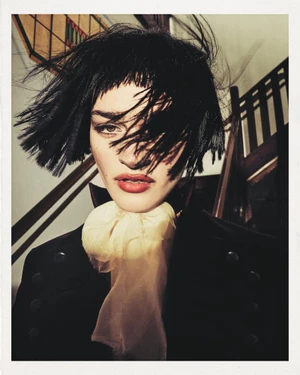 Model with dark short hair and a large cream bow tie wearing a dark jacket inside with stairs behind