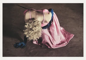 Model with blonde curly hair in a pink dress and blue gloves kneeling on a dark floor