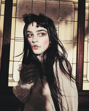 Portrait of a woman with long dark hair, theatrical makeup, and a white blouse standing before a stained glass window.